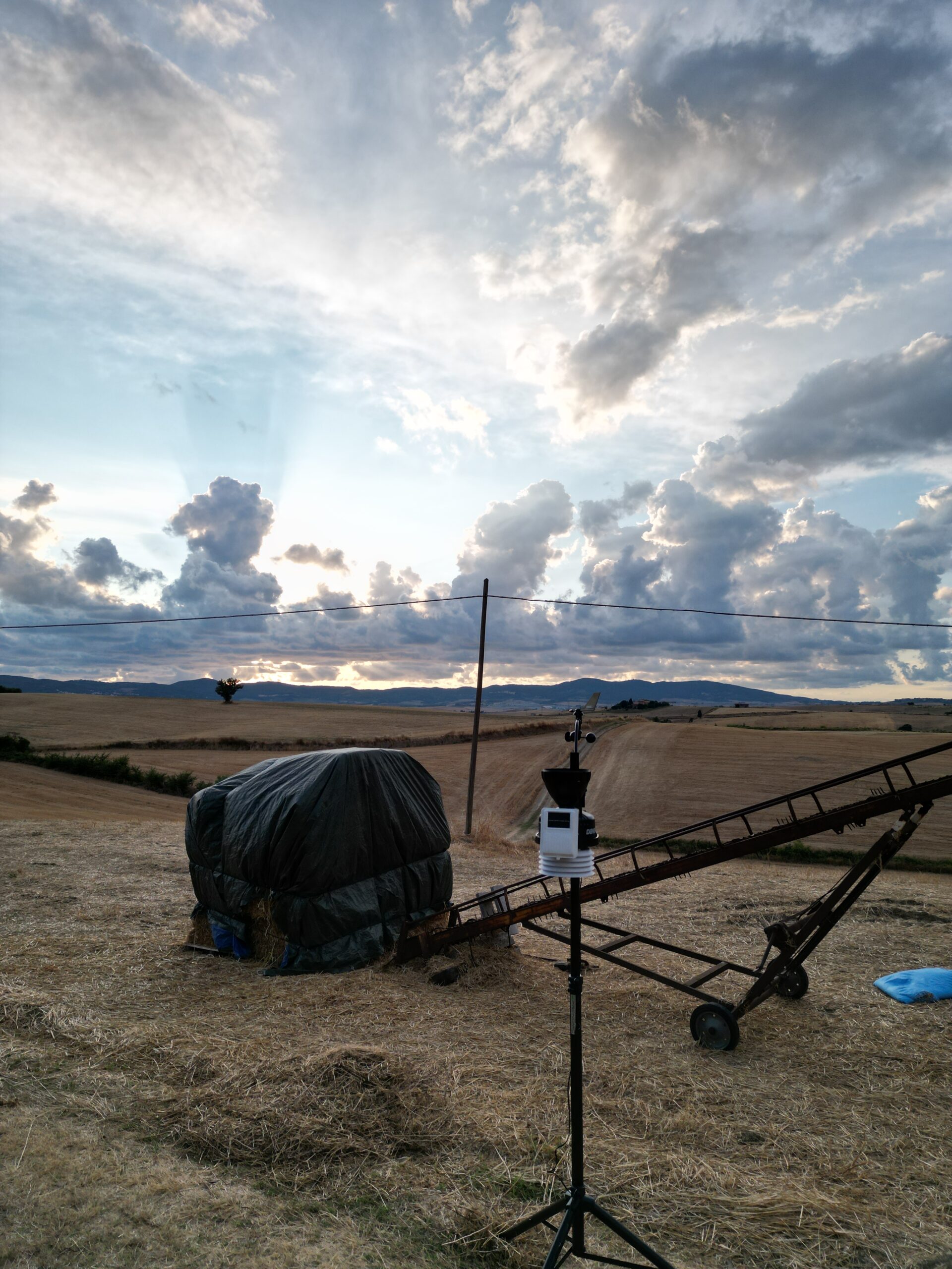 Rural field at harvest with a wrapped hay bale and irrigation equipment under a cloudy sky.